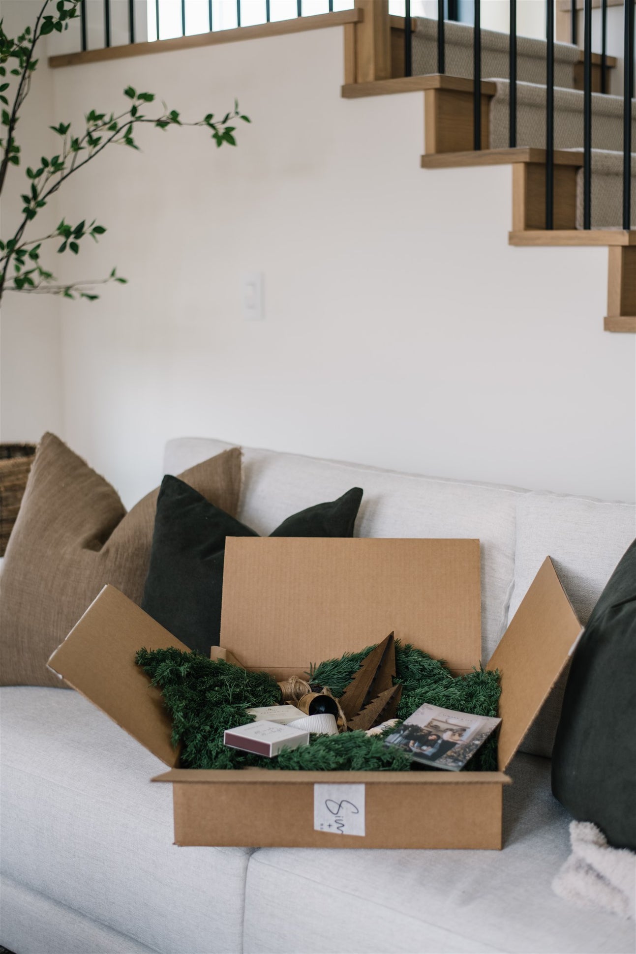 Open cardboard box with greenery and items on a white couch with a staircase in the background