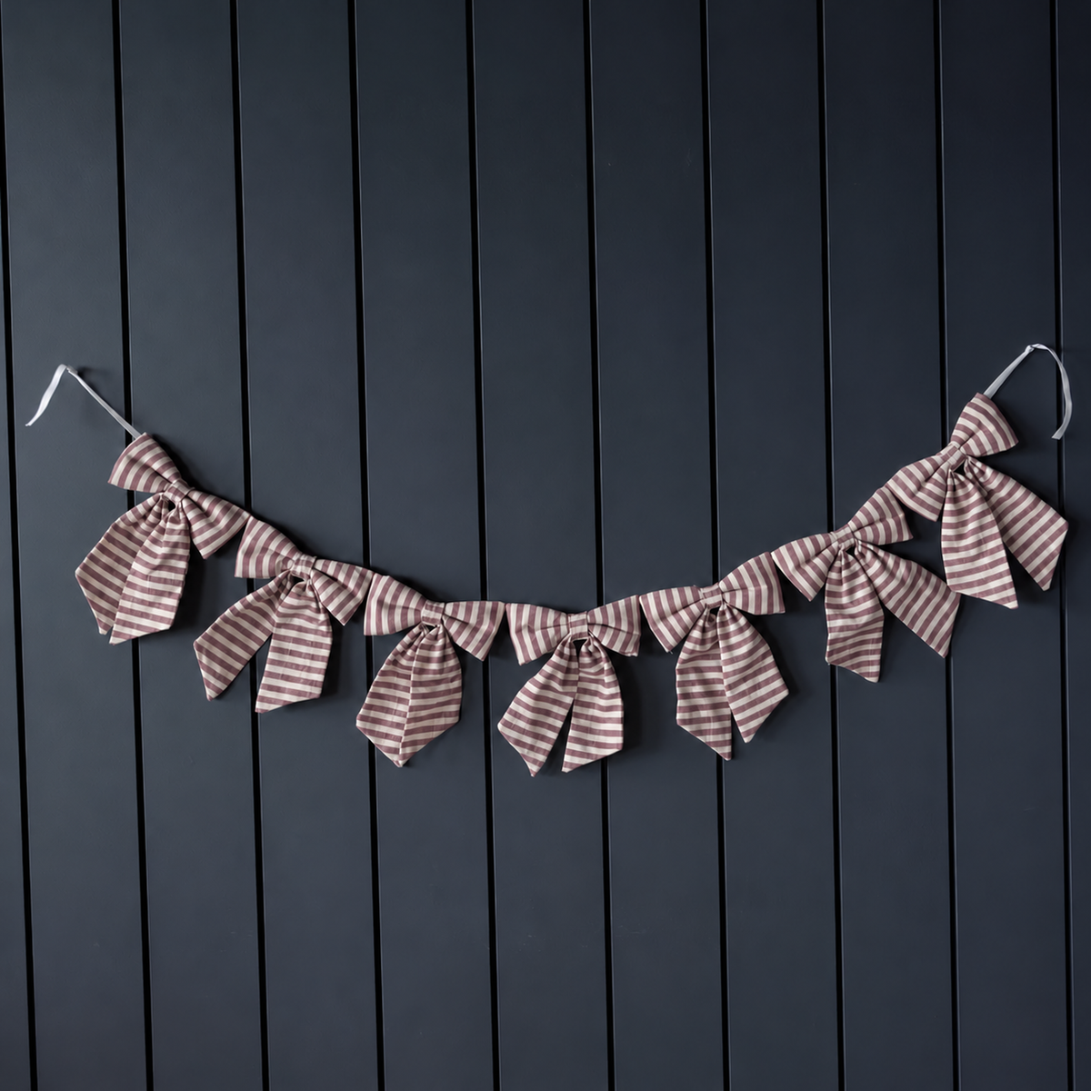 Seven muted cranberry and cream striped cotton bows draped as a garland against a dark panelled wall